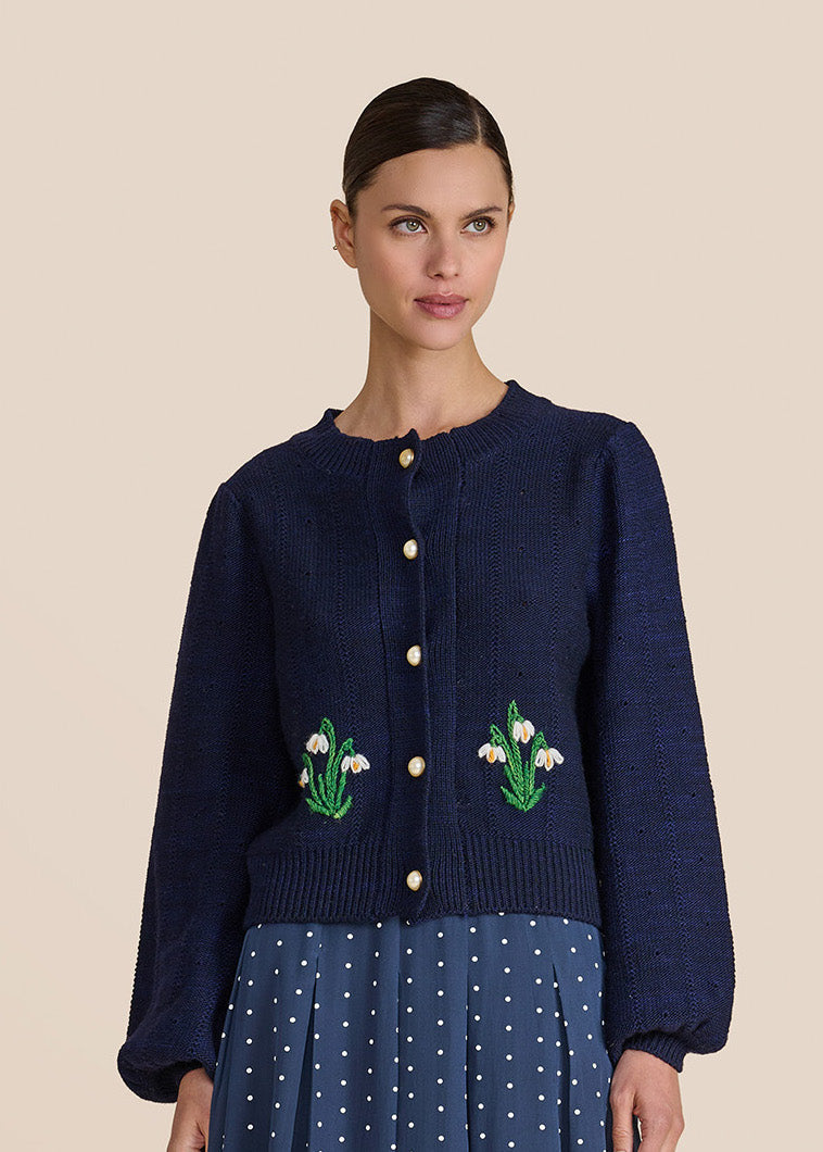 A woman modeling a navy blue knit cardigan with embroidered white and green floral motifs on the front, paired with a blue polka-dot skirt, against a solid beige background.